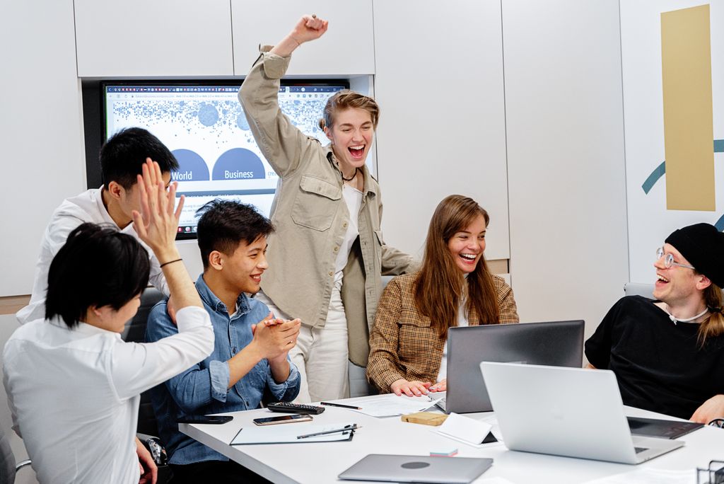 A diverse team enthusiastically celebrating success around laptops during an office meeting.