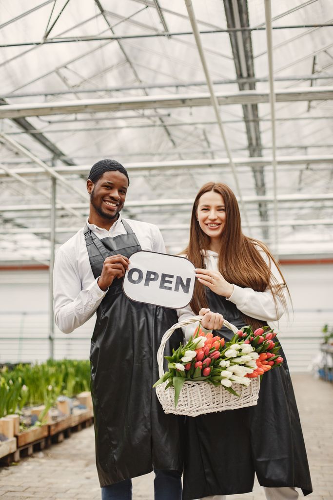 Smiling florists in a greenhouse holding an 'open' sign and a basket of tulips.