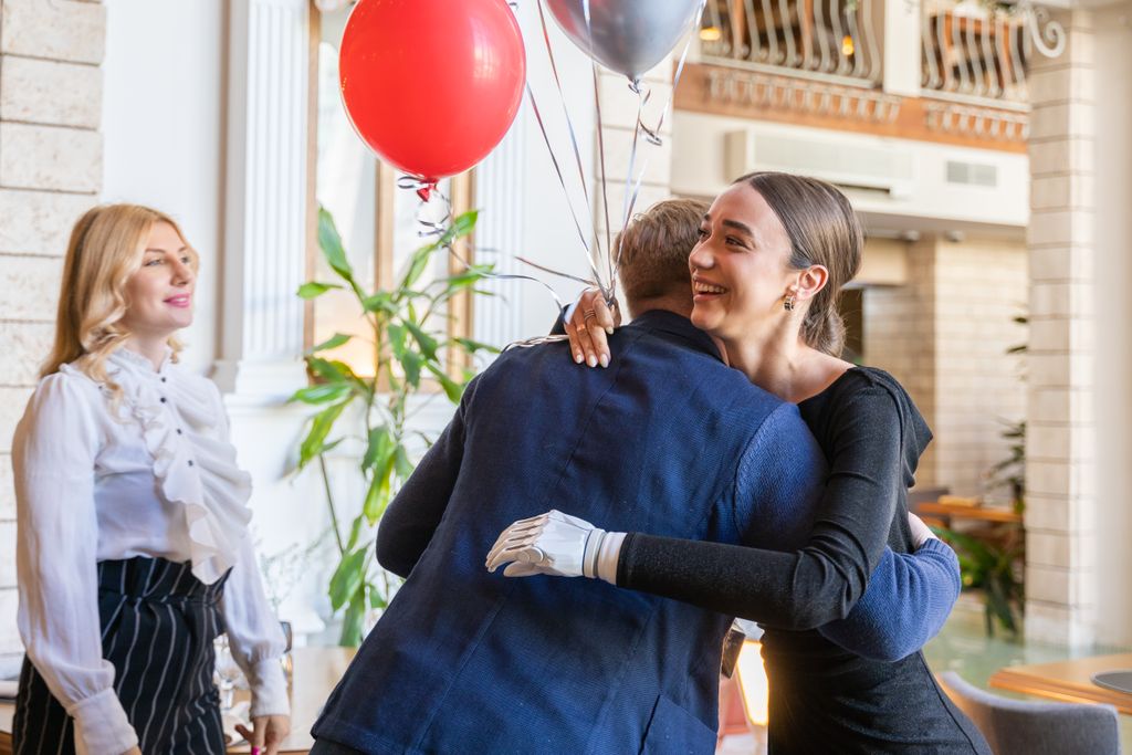 Three people embrace joyfully indoors with festive balloons, celebrating a reunion.